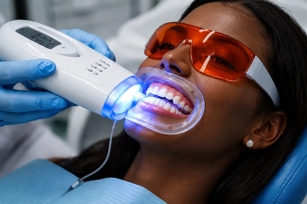 A close-up of a smiling Black woman wearing protective orange eyewear while undergoing a professional teeth whitening treatment with a blue light device